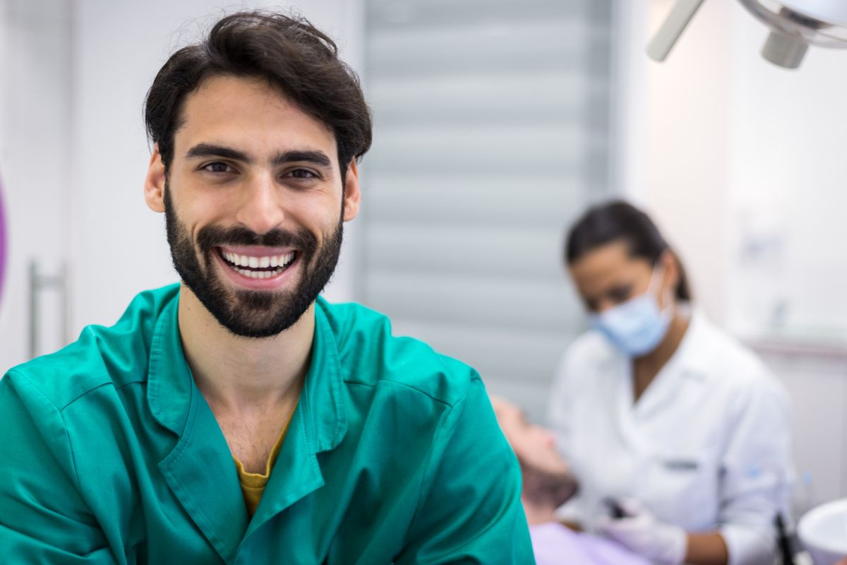 Smiling male dentist in teal scrubs at dental office with colleagues in background.