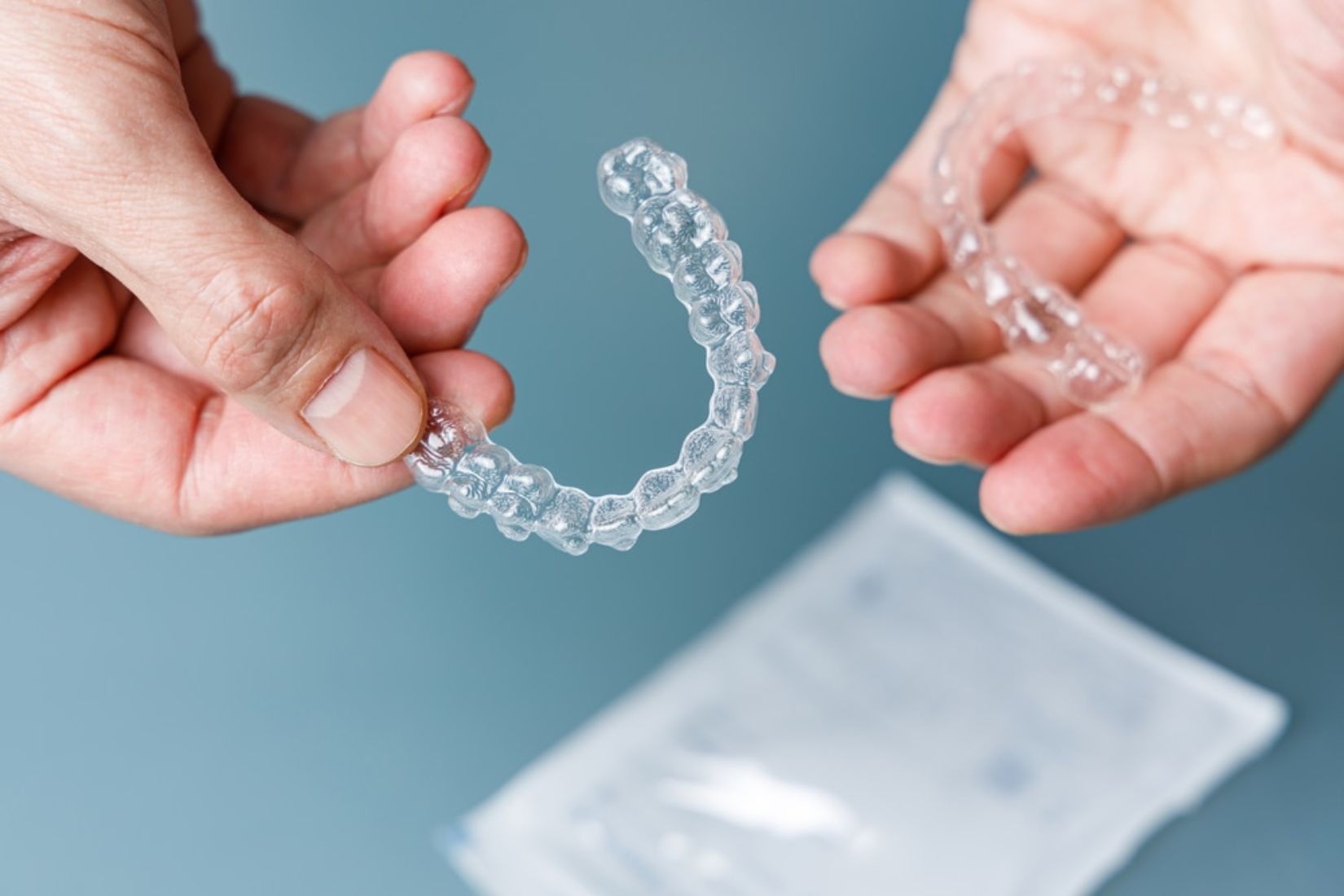 Hands holding clear orthodontic aligner above storage case on blue background.