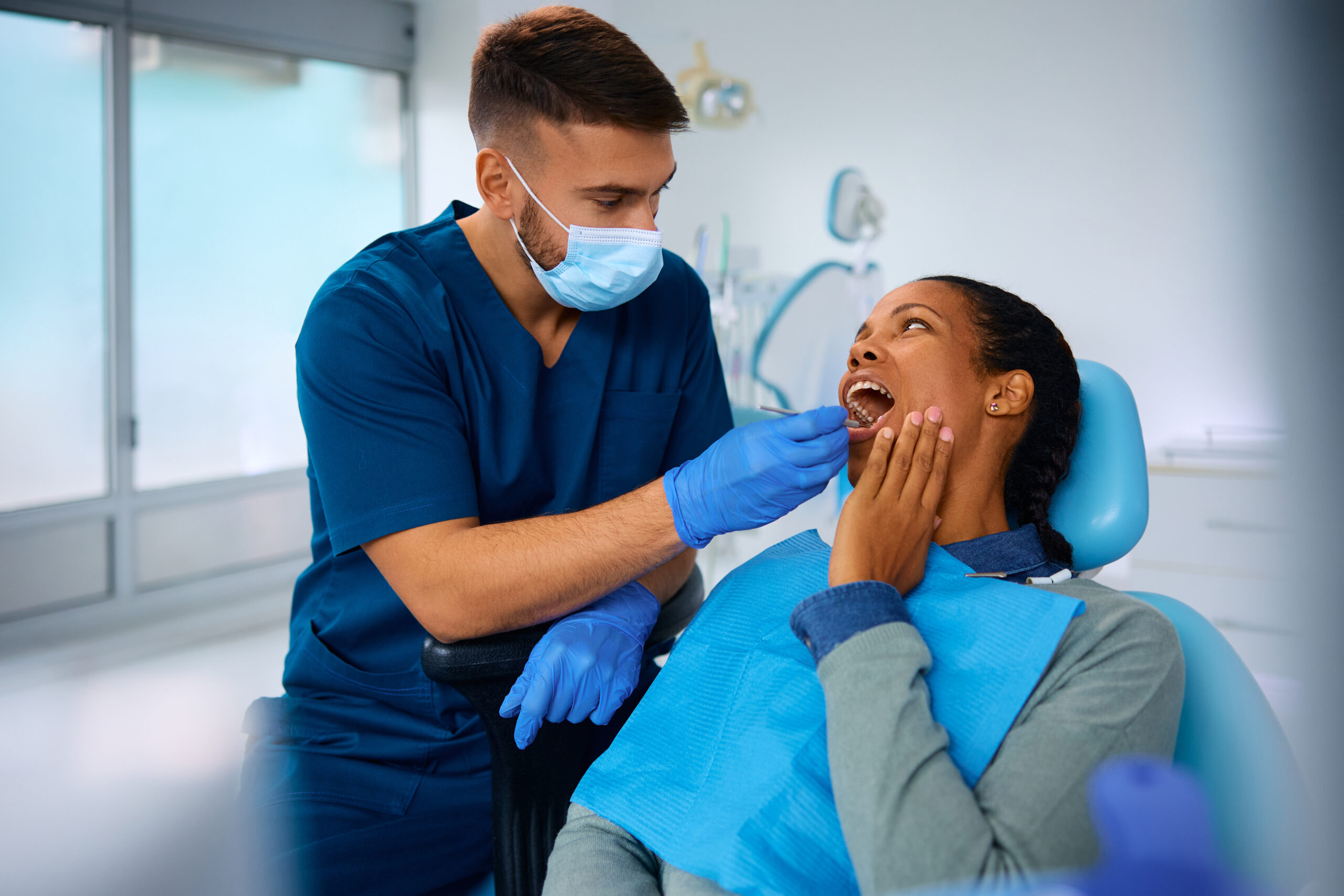 Dentist in blue scrubs examines female patient in dental chair at modern clinic office.