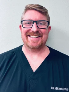 Professional headshot of male dentist wearing glasses and dark scrubs against white background.